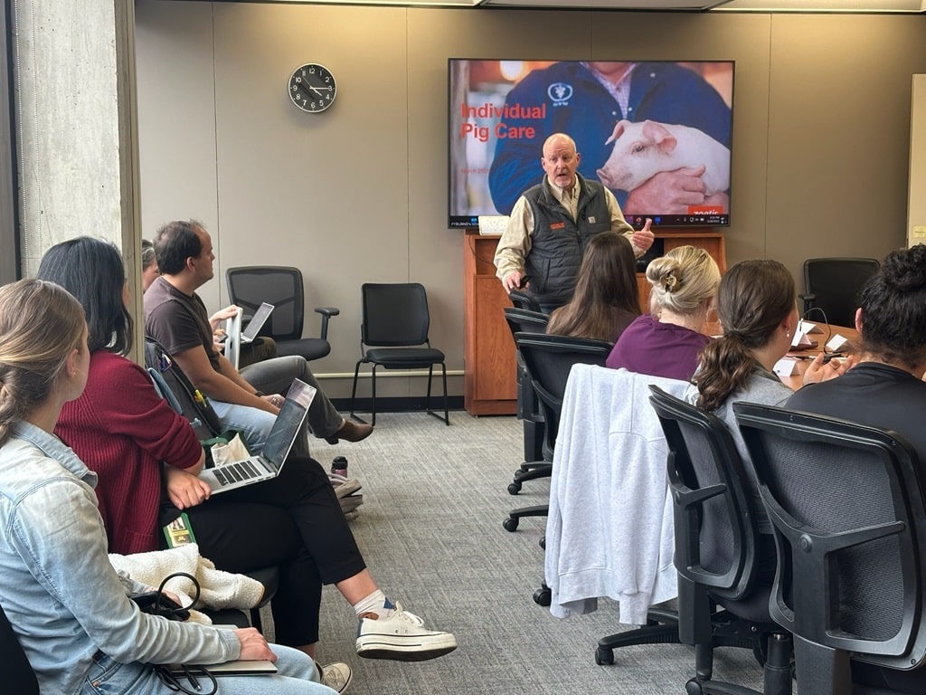 Veterinary instructor speaking with students, surrounded by gestating sows in stalls, during a swine barn walkthrough.